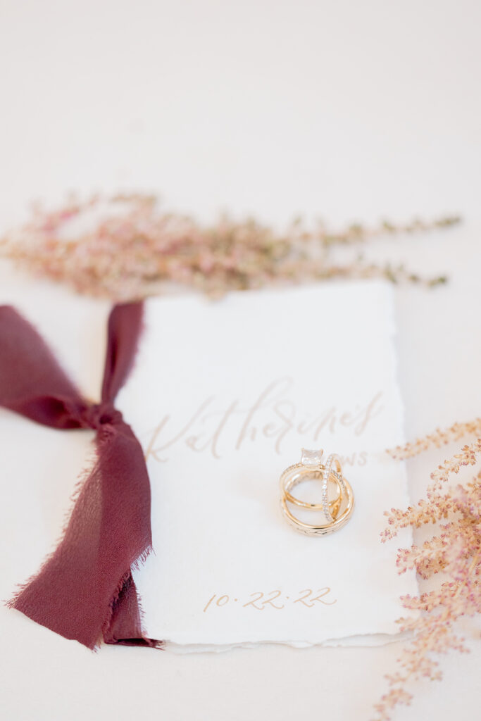 Elegant vow booklet with gold rings, burgundy ribbon, and soft florals on a light background