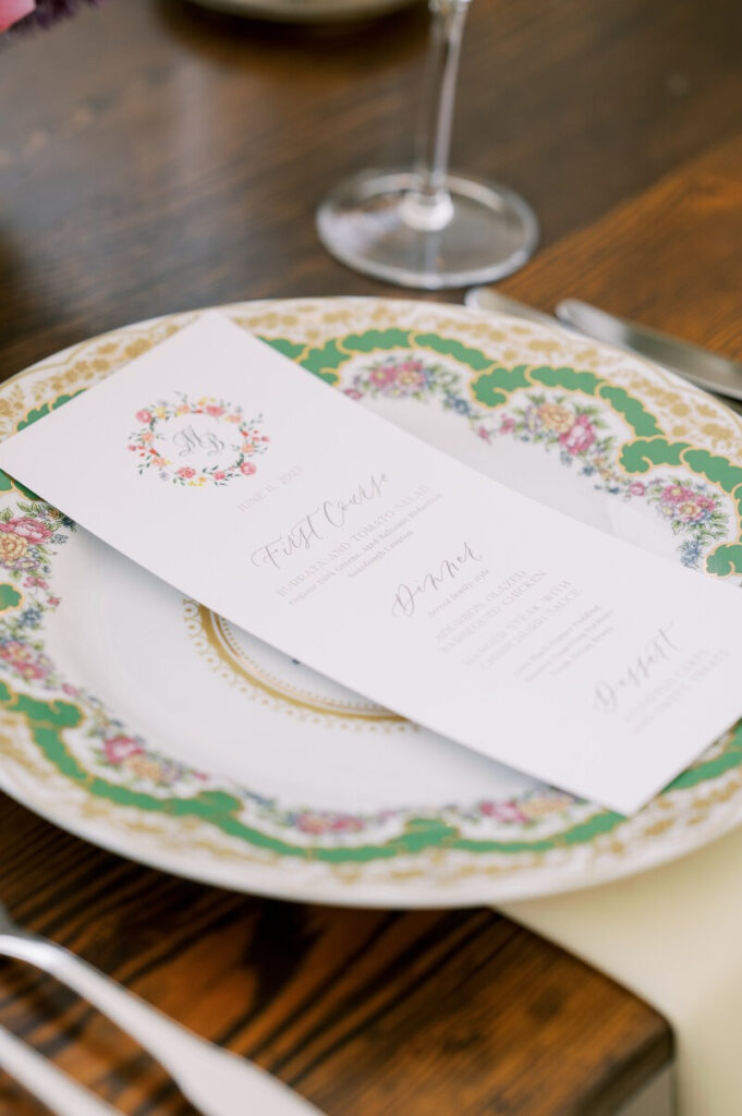 Elegant wedding menu placed on a floral china plate, styled on a wooden table with silverware and glassware.
