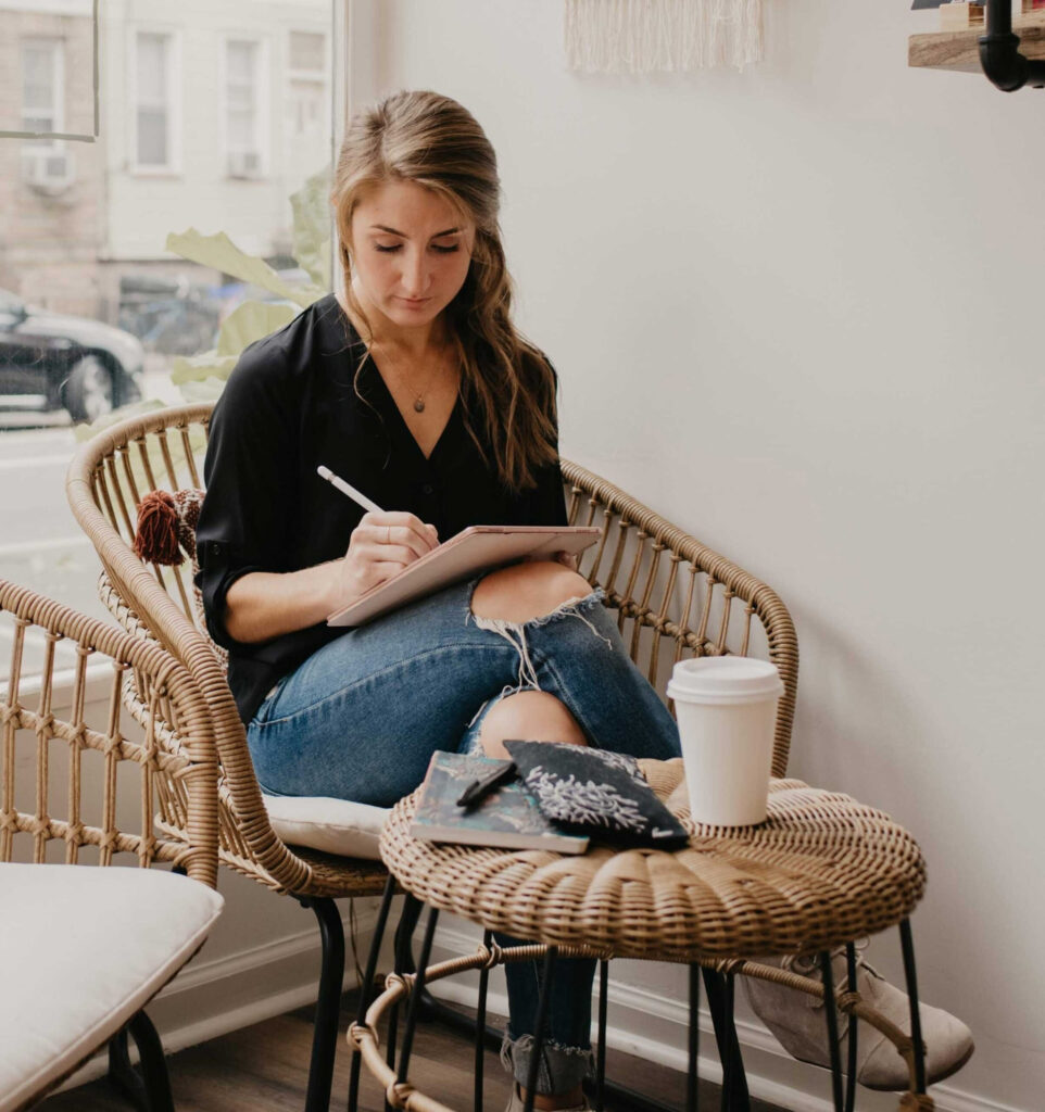 Woman sitting in a wicker chair by a window, writing on a tablet with a stylus, with a coffee cup and notebook on a small table nearby.







