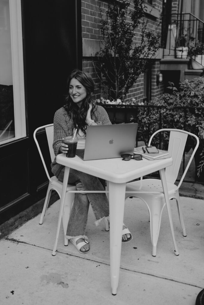 Woman sitting at an outdoor café table with a laptop and iced coffee, smiling while working. This image could represent a creative professional considering the question: should you start a podcast for your wedding business