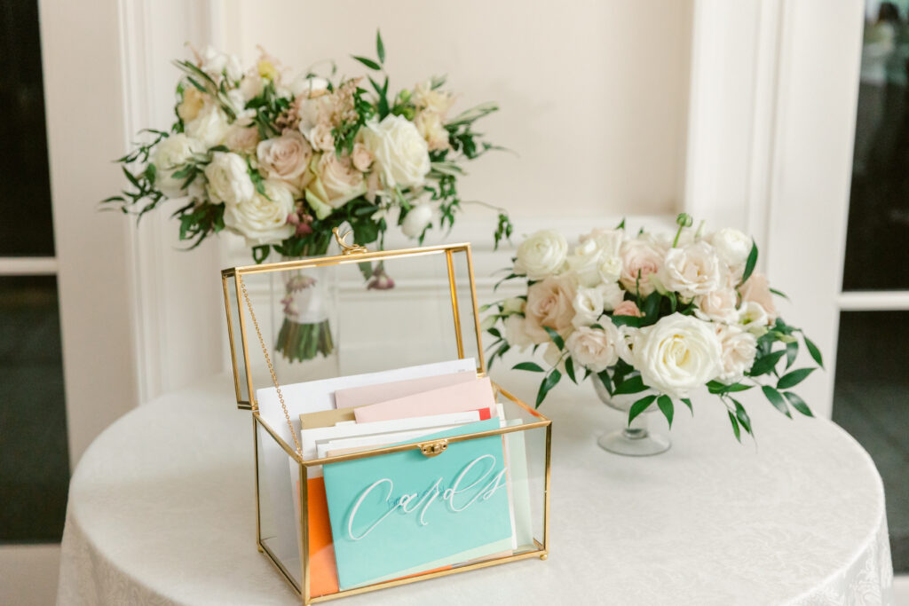 Elegant wedding card box filled with envelopes on a round table, surrounded by white and blush floral arrangements. A fitting visual for vendors asking should you start a podcast for your wedding business to share tips and connect with engaged couples.