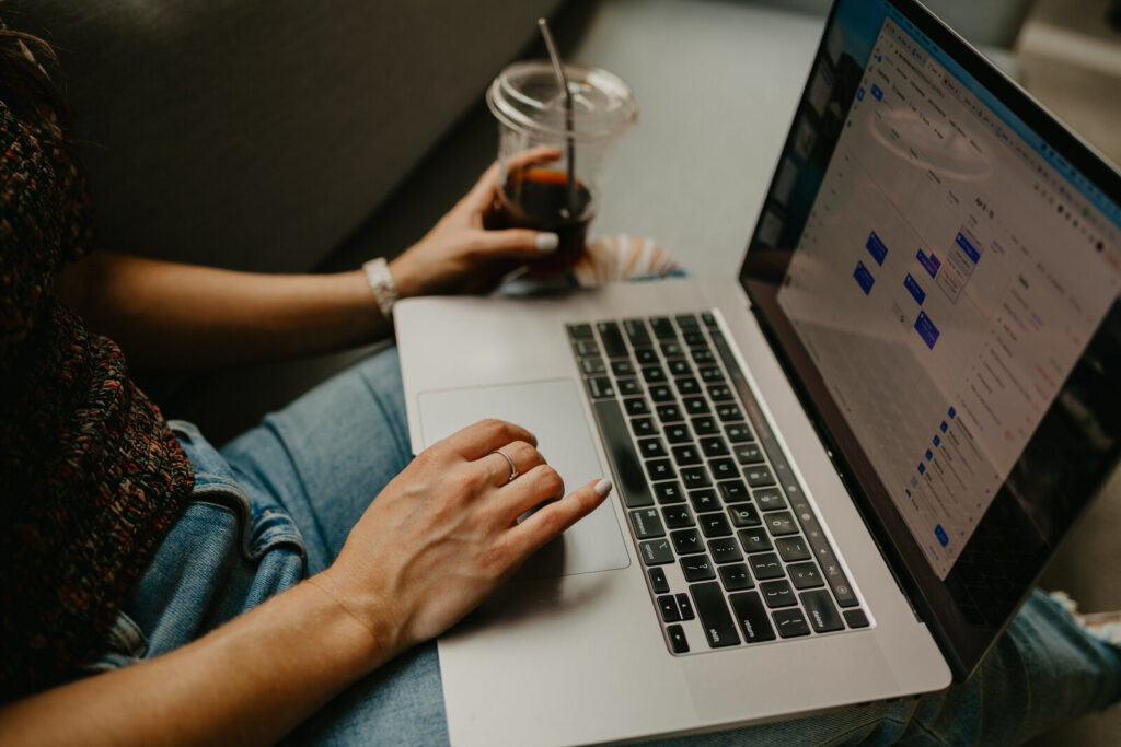 Person sitting on a couch using a laptop with a scheduling app open, holding an iced drink.