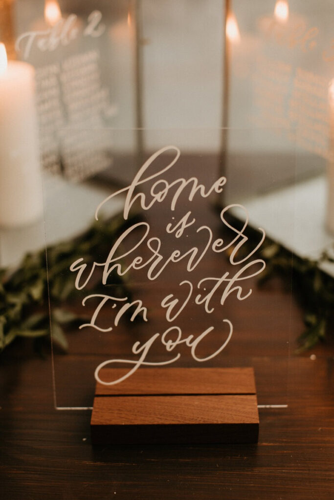Acrylic sign with handwritten calligraphy reading "home is wherever I'm with you," surrounded by candles.