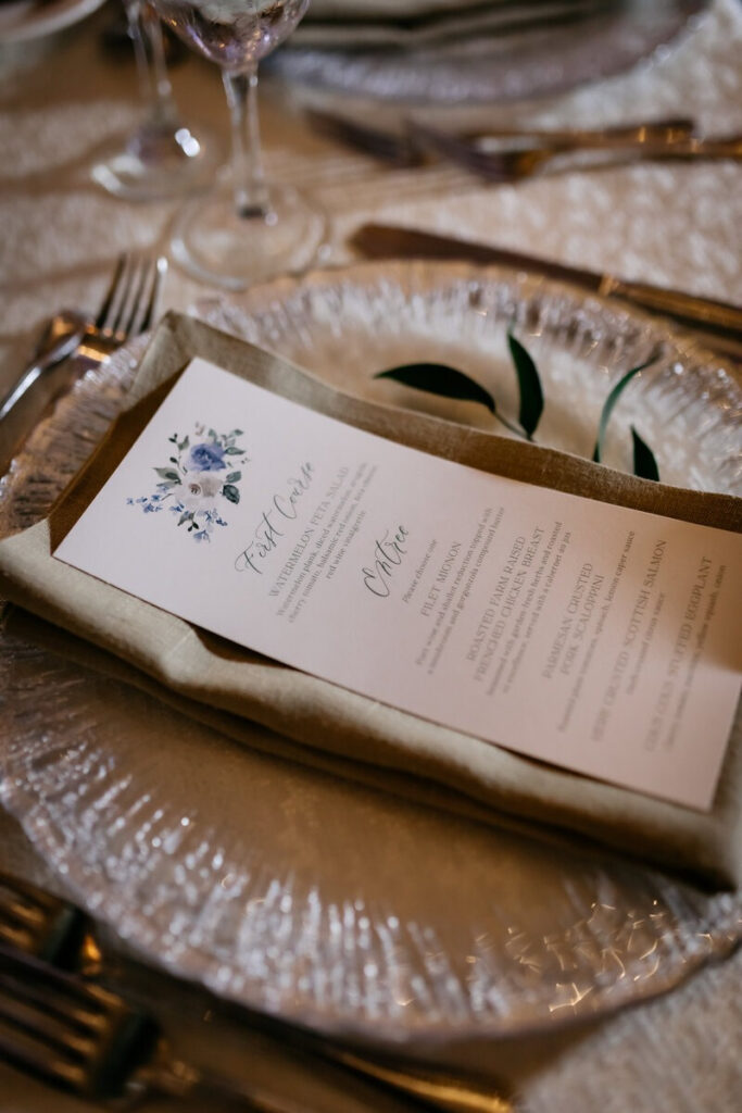 Wedding menu with floral design placed on a linen napkin and glass charger at a formal table setting.